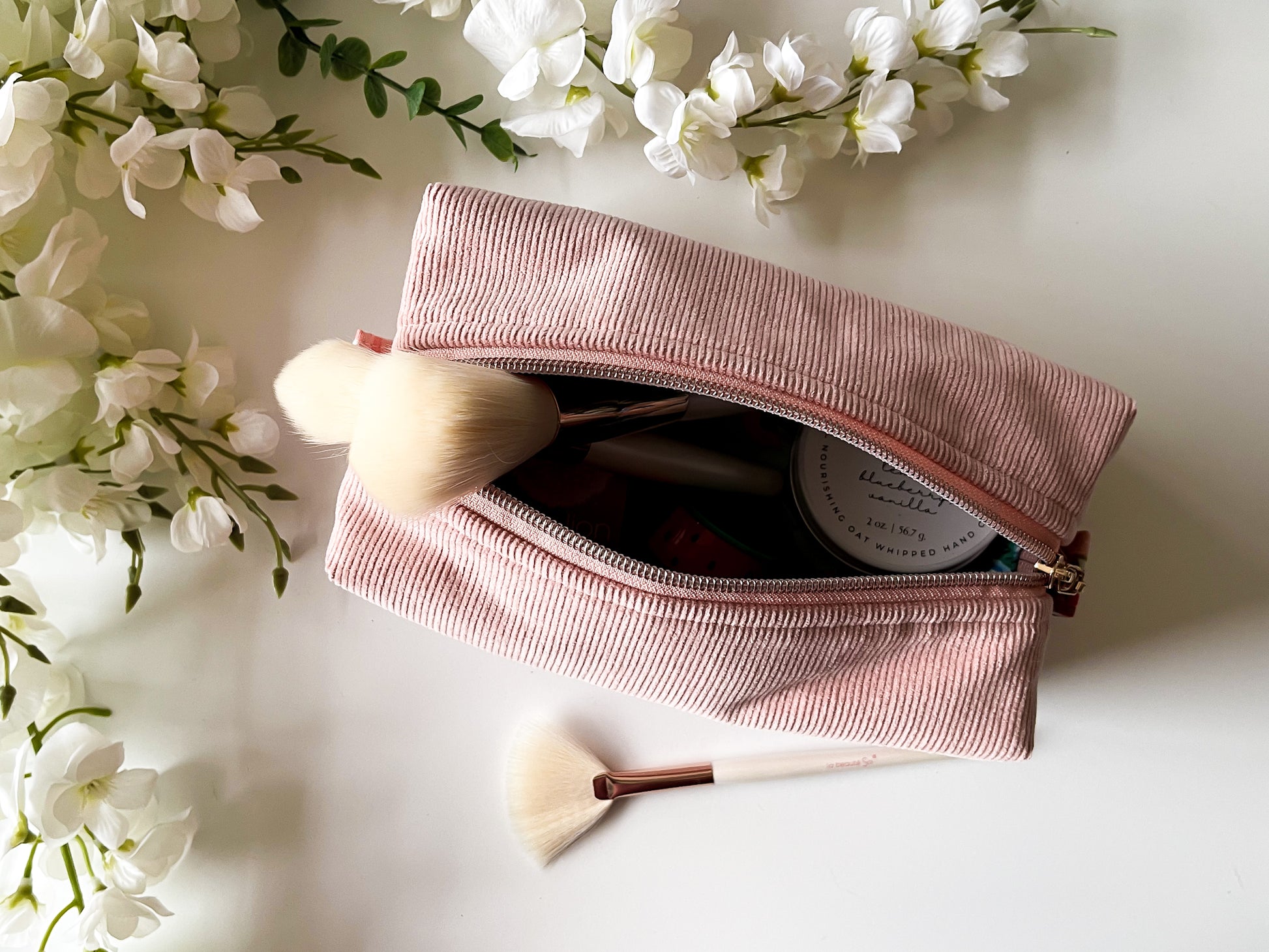 Pink makeup pouch with brushes and a product, surrounded by white flowers on a light background