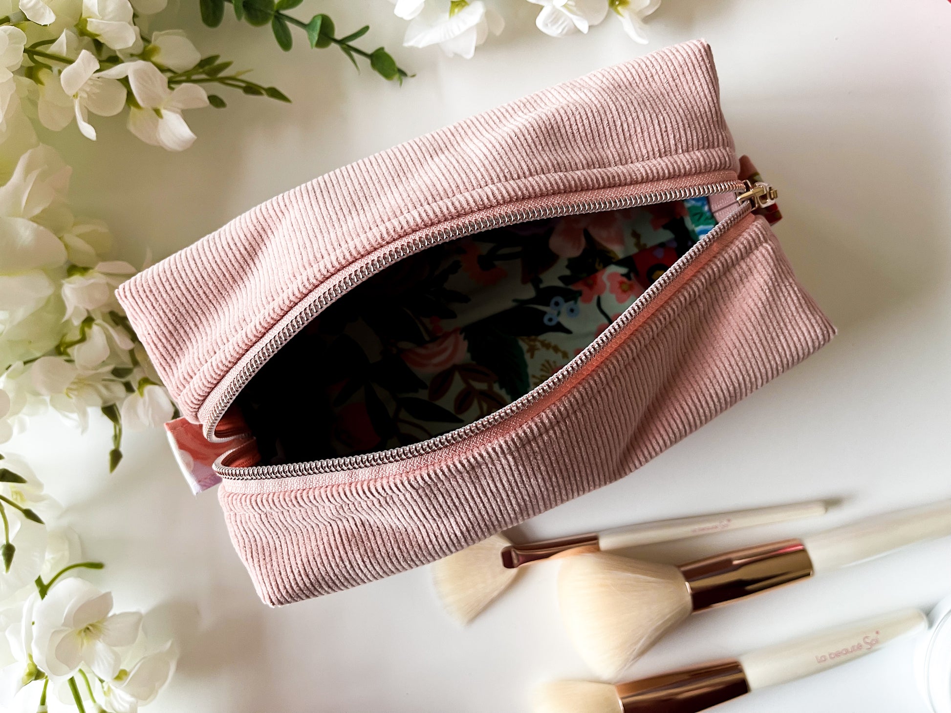 Pink makeup pouch with floral design, surrounded by white flowers and makeup brushes on a light surface.