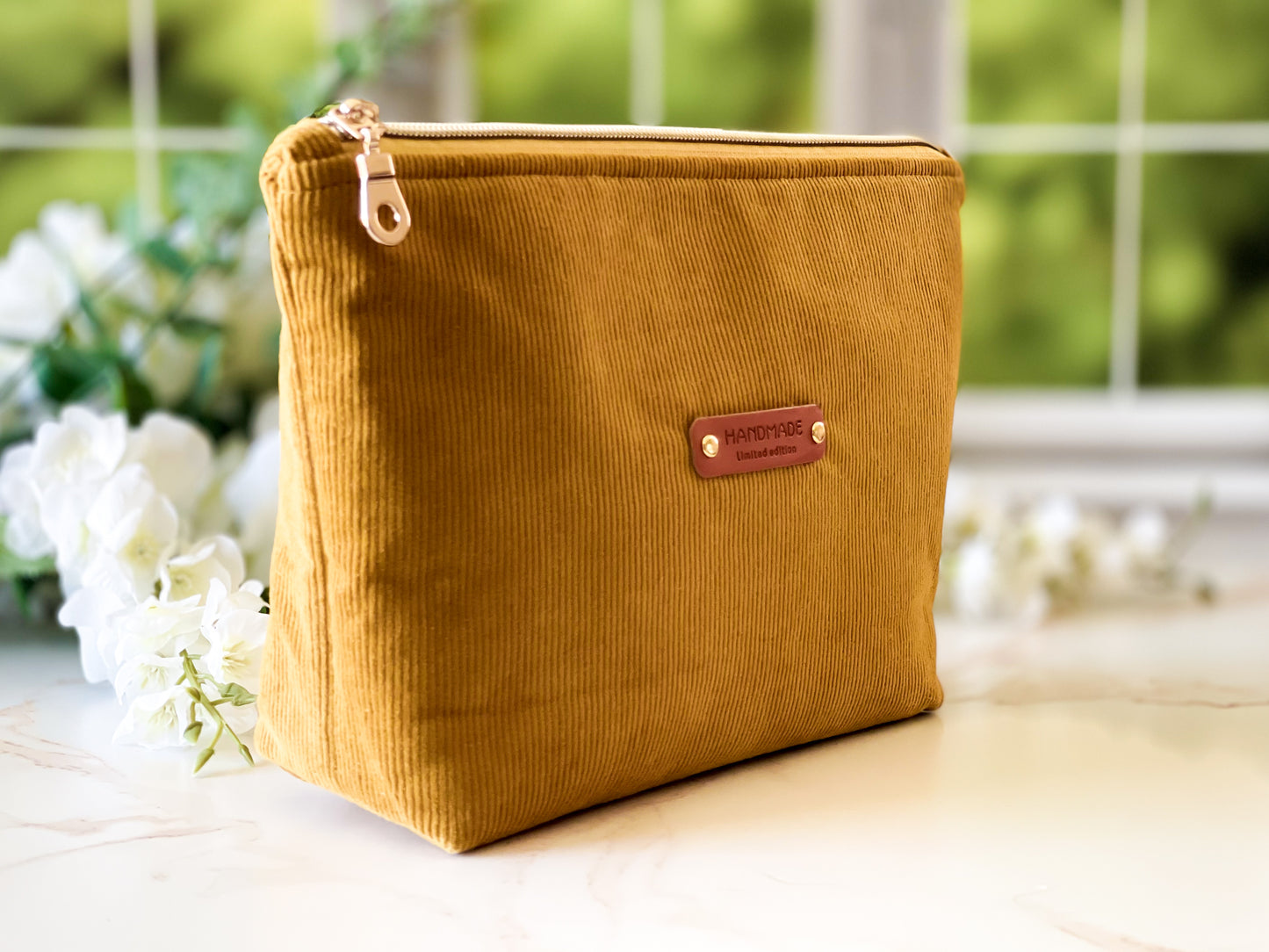 Yellow corduroy pouch with a brown leather patch on a light surface with blurred greenery in the background