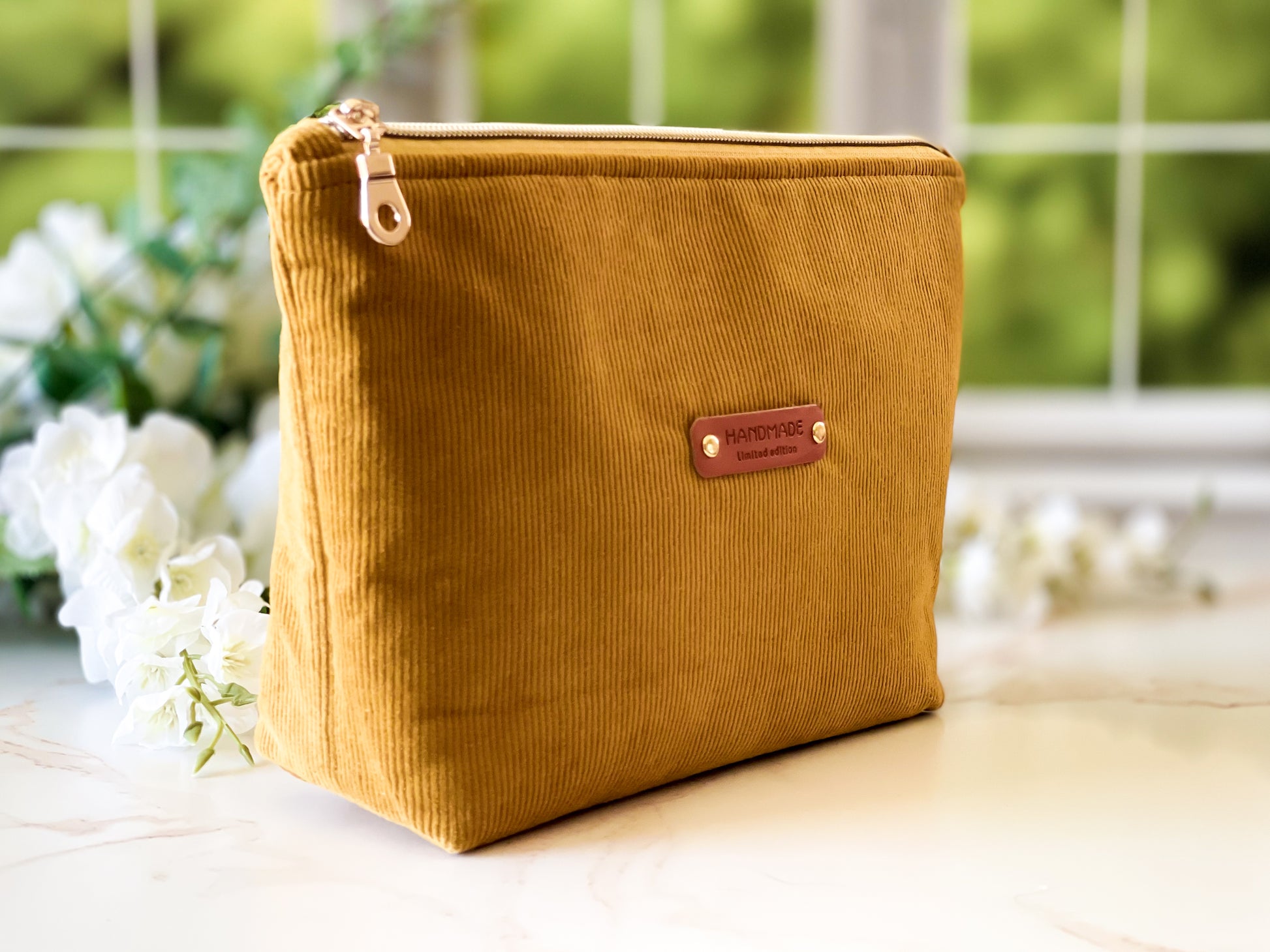 Yellow corduroy pouch with a brown leather patch on a light surface with blurred greenery in the background