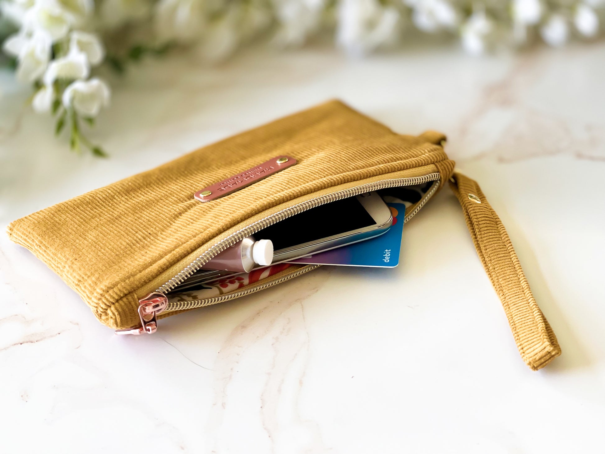 Yellow clutch purse with items on a light surface with blurred flowers in the background