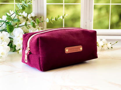 Merlot Corduroy Boxy Bag - Maroon toiletry bag with gold zipper and brand name on a light surface with greenery in the background