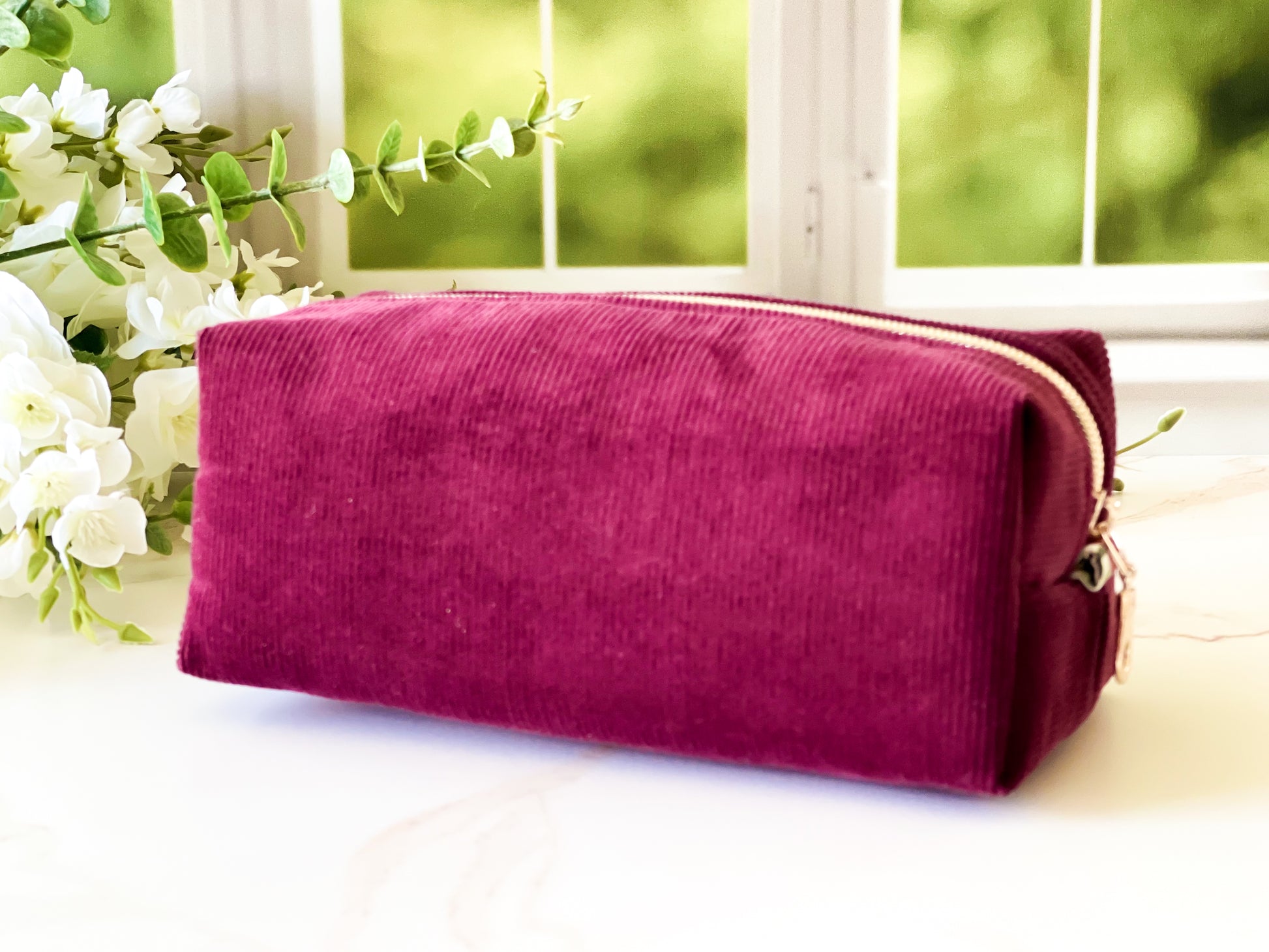 Merlot Corduroy Boxy Bag Maroon pouch with gold zipper on a light surface with greenery in the background
